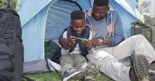 アフリカ系アメリカ人の父親と息子はテントで時間を楽しむ 彼らは屋外キャンプの冒険の間にスマートフォンと瞬間を共有しています スローモーション — ストック動画