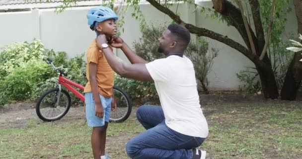 非裔美国人的父亲帮助一个儿子在户外带头盔 他们准备在一个阳光灿烂 绿色的后院慢吞吞地骑自行车 — 图库视频影像