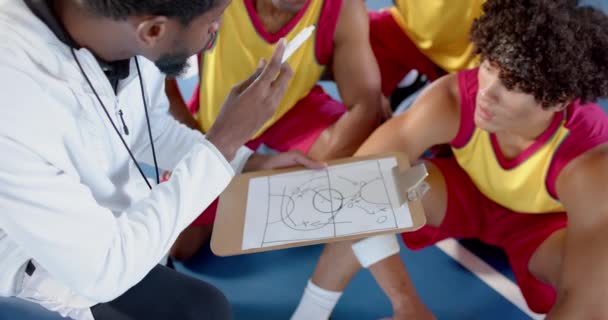 アフリカ系アメリカ人のコーチがスポーツ施設で若い選手たちと戦略を話し合っている チームはバスケットボールのトレーニングセッション中に彼らのゲームプランを改善することに焦点を当てています — ストック動画