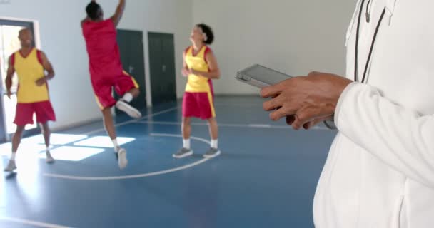 コピースペース付きのバスケットボールコートでタブレットを保持するコーチ 彼は屋内スポーツ施設で練習セッション中にプレーヤーを観察し スローモーション — ストック動画