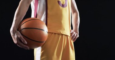 Basketbol formalı bayan basketbolcu siyah arka planda bir top tutuyor. Topa odaklanıldığında spor teması ve rekabetçi ruh, yavaş çekim vurgulanıyor..