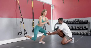 Genç beyaz bir kadın spor salonunda Afro-Amerikalı bir adamla antrenman yapıyor. İyi donanımlı bir çalışma alanında, ağır çekimde fitness hedeflerine odaklanırlar..