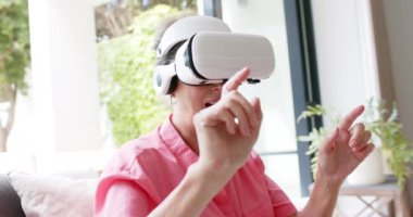Beyaz kadın sanal gerçekliği kendi evinde keşfediyor. Parlak bir oturma odası ortamında, ağır çekimde VR teknolojisiyle meşgul..