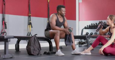 Formda Afro-Amerikalı bir erkek ve beyaz bir kadın spor salonunda dinleniyor. Antrenman sonrası su içiyorlar ve iyileşiyorlar. Sağlıklı bir yaşam tarzı sergiliyorlar. Yavaş çekimde..