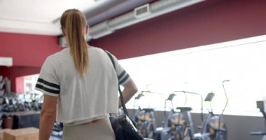 Spor salonundaki genç beyaz kadın, fotokopi odası olan. Egzersiz için hazırlanıyor. Etrafı egzersiz ekipmanlarıyla çevrili. Yavaş çekim..