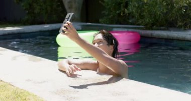 Asyalı ergen çocuk akıllı telefon kullanarak havuz başında güneşli bir günün tadını çıkarıyor. Telefonuyla anı yakalıyor, evde eğlence ve teknolojinin bir karışımını vurguluyor, ağır çekimde..