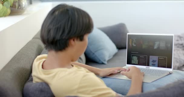 아시아 소년은 집에서 자신의 노트북을 검색합니다 온라인 학습에 편안하게 소파에 — 비디오