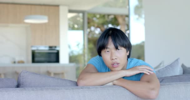 有思想的少年亚洲男孩把下巴放在家里沙发上的胳膊上 他的表情暗示着在明亮的 现代的 慢动作的生活空间里沉思或厌烦 — 图库视频影像