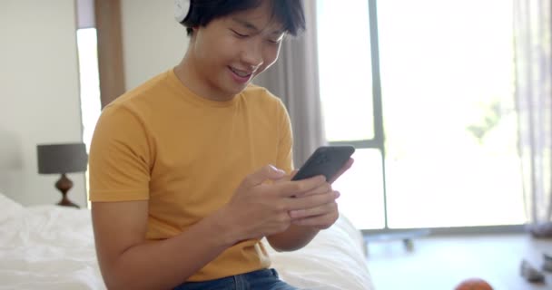 十几岁的亚洲男孩喜欢在家里用手机听音乐 当他在卧室里放松下来时 他的微笑暗示他有片刻的闲暇 慢动作 — 图库视频影像