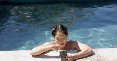 Asyalı genç çocuk havuz kenarında fotokopi odasıyla güneşli bir günün tadını çıkarıyor. Gevşeyip yavaş çekimde kontrol ederken boş zamanı yakalıyor..