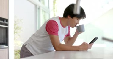 Asyalı ergen çocuk kahvesini yudumluyor ve telefonunu mutfakta kontrol ediyor. Parlak, modern ve ağır çekimde sakin bir anın tadını çıkarıyor..