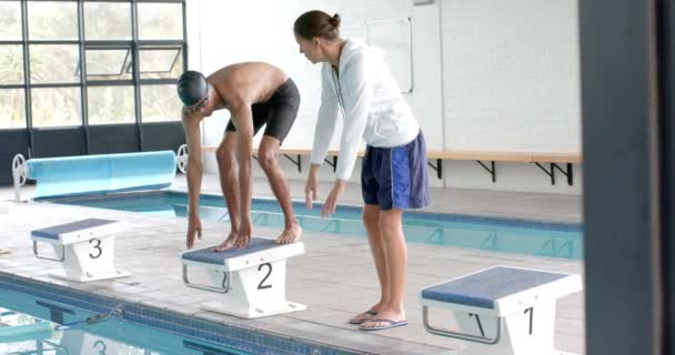游泳教练在游泳池边指导一位年轻的伯利兹男子运动员游泳 他们在一个室内游泳设施 专注于潜水技术 慢动作 — 图库视频影像