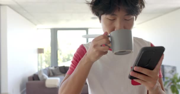 Asiatischer Junge Checkt Hause Sein Handy Balanciert Einen Becher Einer — Stockvideo