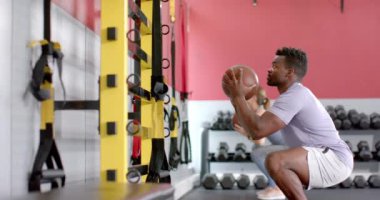 Spor salonunda tıp topuyla egzersiz yapan Afro-Amerikalı bir adam ve beyaz bir kadın. Vücut geliştirme rutinlerine odaklanmışlar. Güç eğitimi için ekipman kullanıyorlar. Ağır çekim..