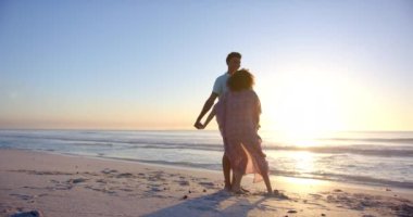 Çift ırklı çift gün batımında sahilde eğlenceli bir yürüyüş yapmaktan hoşlanır. Pembe elbiseli genç kadın beyaz gömlekli adamla gülüyor, ikisi de kumda yalın ayak, ağır çekimde..