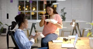 Genç Afrikalı Amerikalı kadın ofiste melez bir kadınla iş yemeği yiyor. Çalışma alanlarında rahat bir yemek molası verirler. Dostça bir atmosferi teşvik ederler. Ağır çekimde..