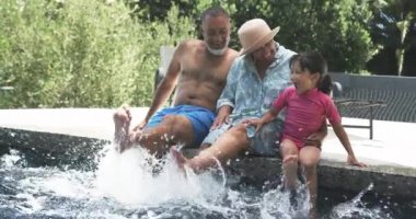 Torunlarıyla birlikte çift ırklı büyükbabalar havuz kenarında su sıçratmaktan hoşlanırlar. 50 yaşındaki adam ve 51 yaşındaki kadın güneşli bir günde 5 yaşındaki çocuklarıyla neşeli bir anı paylaşıyorlar..