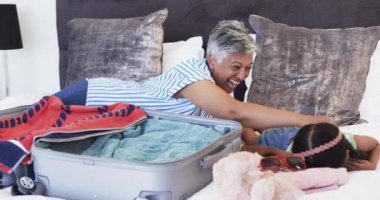 Kısa gri saçlı melez bir kadın bavulunu toplarken gülüyor. Çift ırklı bir kız, oyuncaklarla ve pembe gözlüklerle çevrili, yatakta uzanıyor..