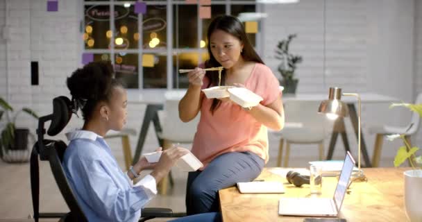 年轻的非洲裔美国女人在办公室和一个嘴上的女人共进午餐 他们在自己的工作空间享受轻松的晚餐休息 营造出友好的氛围和慢动作 — 图库视频影像