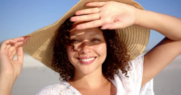 かわいい髪の若い女性は 晴れたビーチで笑顔を浮かべ 手で目を守りました 彼女は日光の中で喜びを放射する広いレンズの帽子と白い衣装を着ています — ストック動画