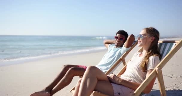 年轻的白人男子和年轻的白人女子在海边的椅子上放松 两人都戴着太阳镜 享受着阳光明媚的海滩氛围 蓝天晴朗 动作缓慢 — 图库视频影像