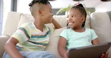 Afro-Amerikalı erkek ve kız kardeş evde bir tabletle neşeli bir anı paylaşıyorlar. Adam çizgili bir gömlek giyiyor ve kızın saçında pembe bir fiyonk var..