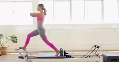 Beyaz bir kadın bir fitness stüdyosunda pilates reformcusuna saldırıyor. Egzersiz rutini, parlak, havadar ve ağır çekimde güç ve esnekliği vurguluyor..
