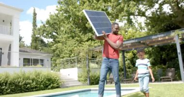 Afro-Amerikalı bir adam güneş paneli tutuyor, evindeki güneşli arka bahçeye bakan bir çocuk var. Çocuğa güneş teknolojisini öğreten adamla birlikte yenilenebilir enerjiyi araştırıyorlar..