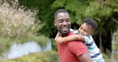 Afrikalı Amerikalı baba, evdeki güneşli bir parkta, fotokopi odasıyla oğlunu sırtında taşıyor. İkisi de gülümsüyor, açık havada neşeli ve sevecen bir anın tadını çıkarıyor, ağır çekimde..