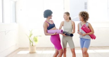 Üç kadın gülümsüyor ve yoga minderlerini parlak bir odada tutuyorlar. Soldaki melez kadın, merkezdeki beyaz kadın ve sağdaki melez kadın, spor sonrası neşeli bir anı paylaşıyorlar..