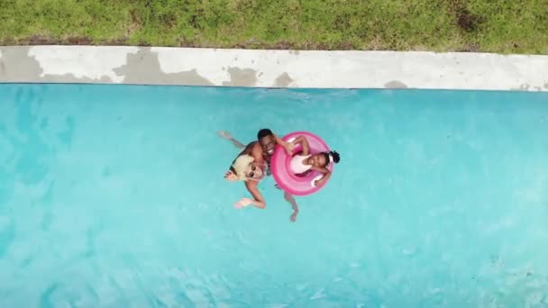一个非裔美国家庭在家里的游泳池里的一个粉色充气环上漂浮着 他们享受一个阳光明媚的日子 家人以一种保护的方式拥抱着他们 — 图库视频影像