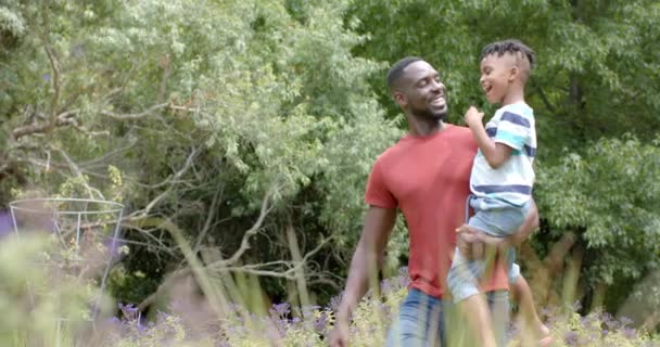 Ojciec Afroamerykanów Niesie Syna Oboje Uśmiechnięci Bujnym Ogrodzie Domu Przestrzenią — Wideo stockowe