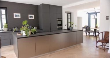 Modern bir mutfakta parlak karanlık dolaplar ve merkezi bir ada bulunur. Doğal ışık geniş pencerelerdeki boşluğu dolduruyor, temiz, çağdaş tasarımı vurguluyor, yavaş çekim.