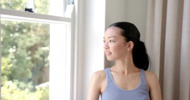 Neşeli bir gülüşü olan melez bir kadın kopya alanı olan bir pencerenin yanında duruyor. Saçları arkadan bağlanmış, mavi bir atlet giyiyor, odayı aydınlatıyor, ağır çekimde..