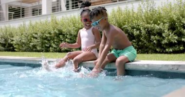 Afro-Amerikalı kız ve erkek kardeşler evlerinde havuz başında eğleniyorlar, ayaklarıyla su sıçratıyorlar. Kadın pembe mayo ve kırmızı güneş gözlüğü takıyor; erkek yeşil şort ve mavi gözlükler giyiyor, yavaş çekim..
