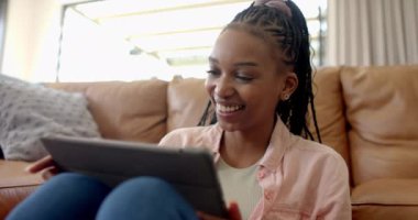 Evde tablet kullanan genç bir Afrikalı Amerikalı kadın. Saçlarını ördü, pembe bir gömleği var ve rahat bir şekilde kahverengi koltukta oturuyor, ağır çekimde.