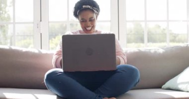 Evdeki kanepede dizüstü bilgisayar kullanarak gülümseyen Afrikalı Amerikalı kadın. Mutlu ve odaklanmış görünüyor. Parlak, güneşli bir odada, ağır çekimde..