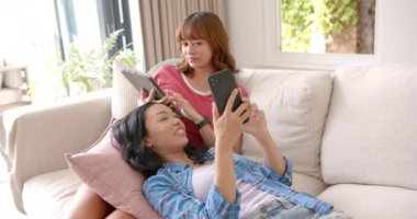 İki melez genç bayan arkadaş kanepede akıllı telefonlara bakarken gülüyorlar. Rastgele giyinerek, rahat bir anın tadını çıkarırlar. Parlak, rahat bir oturma odası ortamında, ağır çekimde..