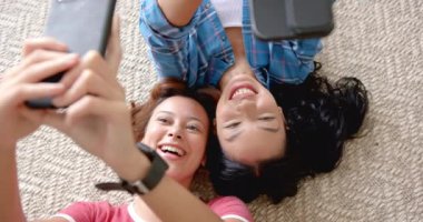 İki melez genç bayan arkadaş yerde uzanıyorlar ve telefonlarıyla selfie çekiyorlar. Neşeli bir şekilde gülümsüyorlar, bir anlık dostluk ve teknolojiyi yakalıyorlar, yavaş çekimde..