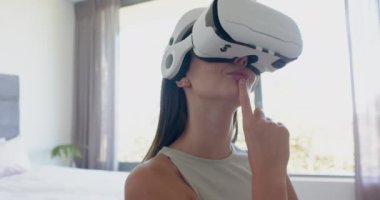 Kafkasyalı, kahverengi saçlı genç bir kız sanal gerçekliğe dalmış, evde bir VR kulaklık takıyor. Etkileşimli bir oyun ya da eğitim simülasyonu ile meşgul ve odaklanmış görünüyor..