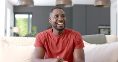 Kırmızı tişörtlü Afro-Amerikalı bir adam video ekranında koltukta rahatça oturuyor. Gülüşü ve rahat duruşu sıradan ve mutlu bir ruh hali olduğunu gösteriyor, yavaş çekim..