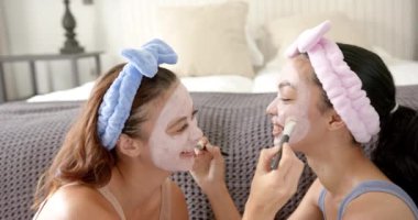 İki çift ırklı genç bayan arkadaş, yüz maskesi ve saç bandı takarak cilt bakımının keyfini çıkarıyorlar. Kameraya gülümsüyorlar, evde bir bağ kurma anı yaratıyorlar, yavaş çekim..