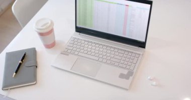 Ofis ortamında beyaz bir masada modern bir iş bilgisayarı. Aksesuarlar arasında bir not defteri, kalem, kahve fincanı ve kablosuz kulaklıklar, profesyonel bir ortam, ağır çekim.