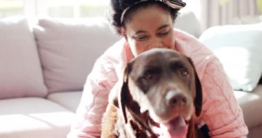 Afro-Amerikalı bir kadın evdeki kanepede kahverengi bir Labrador köpeğine sarılıyor. Sıcak güneş ışığı filtreleri, evcil hayvan ve sahibi arasındaki samimi anın altını çizmek, yavaş çekim.