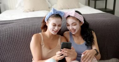 İki melez kız arkadaş, yüz maskeleri ve saç bantlarıyla akıllı telefona gülüyorlar. Birlikte gülüyorlar, akıllı telefona bakıyorlar, rahat bir odada bir yatakta oturuyorlar..