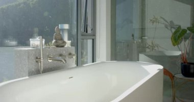 Beyaz küvetli sakin bir banyo büyük bir pencereden doğal ışıkla yıkanır. Buda heykeli ve bitkilerin varlığı rahatlamak için sakin, spa benzeri bir atmosfer izlenimi veriyor..