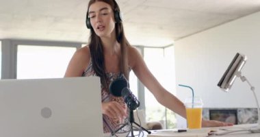 Uzun kahverengi saçlı beyaz bir kız bilgisayar kullanıyor ve kulaklık takıyor. İşine odaklanmış görünüyor. Çevrimiçi öğrenme ya da uzaktan öğrenme ile meşgul. Yanında bir içecekle..
