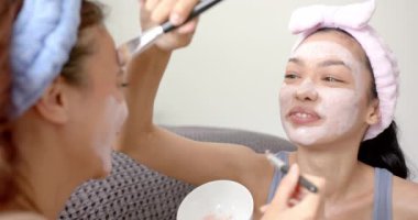 İki melez kız arkadaş yüz maskesi takıyor, biri diğerine yardım ediyor. Saç bandı takan ve elinde fırça tutan biri ile cilt bakımı rutininin tadını çıkarıyorlar..