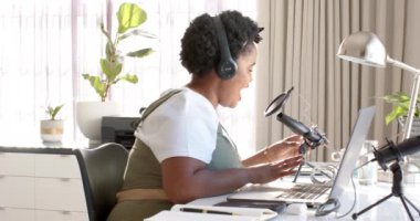 Kulaklıklı Afrikalı Amerikalı bir kadın evdeki bir mikrofona konuşuyor ve bir podcast kaydediyor. İyi aydınlatılmış bir ofis ortamında, ağır çekimde içerik yaratmaya odaklanmış durumda..