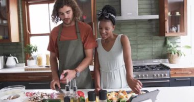 Genç beyaz adam ve Afrikalı Amerikalı kadın modern bir mutfakta birlikte yemek hazırlıyorlar. Ona rehberlik ederken bir yumurtayı kırıyor, ikisi de önlük giyiyor ve taze malzemelerle çevrili..